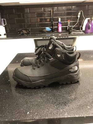 Svarta boots från The North Face - Säljer ett par svarta boots från The North Face med grov sula och snörning. Skorna är tillverkade i slitstarkt syntetmaterial och har logga på sidan samt tungan. Perfekta för höst och vinter med sin robusta design och höga komfort.