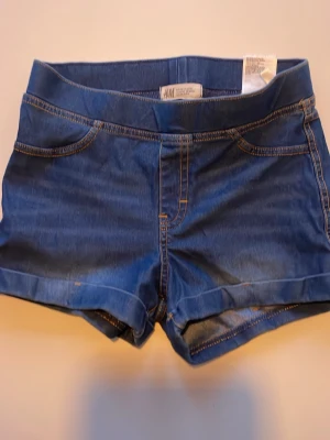 Blå jeansshorts från H&M - Säljer ett par klassiska blå jeansshorts från H&M med normal passform. Shortsen har midwaist och snygga detaljer med kontrastsömmar. Perfekta för varma dagar och har en enkel, tidlös look i stretchigt material 
