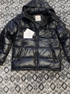 Svart Moncler pufferjacka med huva - Säljer en svart pufferjacka från Moncler med glansig finish och huva. Jackan har dragkedja framtill, två snedställda fickor med dragkedja och en ficka på ärmen med Moncler-logga. Perfekt för kalla dagar och riktigt snygg streetstil.