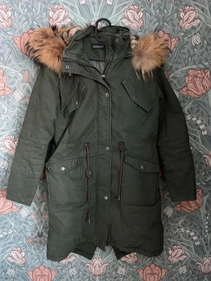 Grön vinterjacka från Rockandblue med pälskant - Säljer en mörkgrön vinterjacka i parkasmodell från Rockandblue med stor huva och fluffig pälskant. Jackan har flera fickor, snygga patchar på ärmen, dragsko i midjan och stängs med dragkedja. Perfekt för kalla dagar och riktigt skön passform. Sömmen har spruckit lite på sidan längst ner, se sista bilden. Är lätt att laga då det går längst sömmen. 