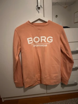 Persikofärgad sweatshirt från Björn Borg - Säljer en snygg persikofärgad sweatshirt från Björn Borg med vit logga framtill. Tröjan har rund halsringning, långa ärmar och är gjord i mjuk bomull. Perfekt för en chill och sportig look.