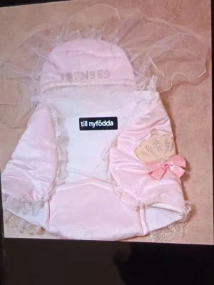 Rosa prinsessväst med spets till nyfödd - Supergullig rosa väst med spetsdetaljer och broderad krona på sidan, perfekt för små prinsessor. Västen har en söt rosett och mjuka tyger som känns lyxiga. Passar till nyfödda och har fina dekorativa inslag som gör den extra speciell.