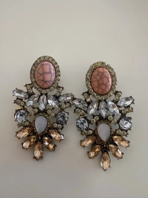 Örhänge oanvända  - Bilderna visar ett par stora, glittriga örhängen med rosa och beige stenar, samt klara kristaller i olika former. Ingen väst syns på bilderna.