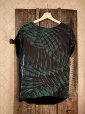 Svart och grön topp från Hugo Boss - Unik topp från Hugo Boss i storlek M med coolt grafiskt mönster i grönt och svart på framsidan och helsvart baksida. Modellen är kortärmad med rund hals och har en loose passform. Materialet är mjukt och känns lyxigt mot huden.