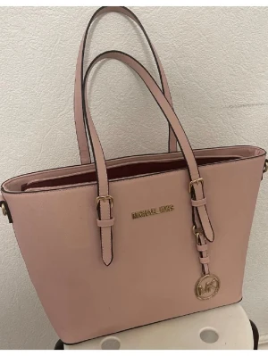Rosa axelväska från Michael Kors - Snygg rosa axelväska från Michael Kors med guldfärgade detaljer och logotyp framtill. Väskan har justerbara axelband, dragkedja upptill och flera fack inuti för smart organisering. Tillverkad i tåligt syntetmaterial och har en klassisk, stilren form.