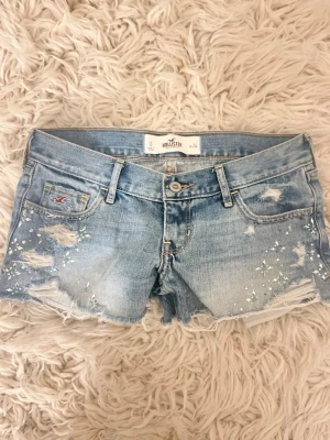 SE ANNAN ANNONS FÖR FRI FRAKT!Ljusblå jeansshorts lågmidjade super lowrise med paljetter Hollister - SKRIV INNAN NI KÖPER! Midja: ca 36 rakt över! Snygga ljusblå jeansshorts från Hollister med slitna detaljer och rå kant. Framtill finns paljetter och små pärlor som ger extra bling. Klassisk femficksmodell med broderad logga på fickan och dekorativa sömmar bak. Perfekta för varma dagar!