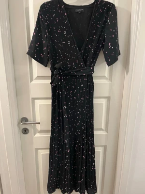 Svart omlottklänning Selected Femme - Svart långklänning från Selected Femme med rosa och vita prickar, omlottmodell och knytband i midjan. Klänningen har korta ärmar och ett plisserat tyg som ger en snygg struktur. Perfekt för dig som gillar en cool och feminin stil.