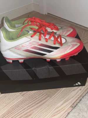 Adidas fotbollsskor f50s.       - Säljer ett par Adidas fotbollsskor med snygg design i vitt, rött och svarta detaljer. Skorna har röda snören, grön insida och platt sula med dobbar för gräsplan. Tillverkade i syntetmaterial och har en rund tå. Perfekt för dig som vill sticka ut på planen!     Inte minsta trasiga men är lite smutsiga