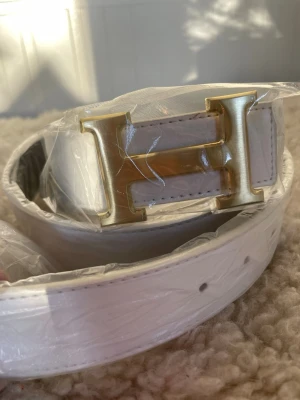 Vit bälte med guldspänne - Snyggt vitt bälte med ett stort, stilrent H-format spänne i guld. Bältet har en slät och lätt texturerad yta med svarta detaljer på insidan. Perfekt för att lyfta din outfit med en lyxig touch och passar till både jeans och kostymbyxor.