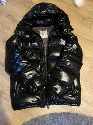 Svart pufferjacka från Moncler - Säljer en svart, glansig pufferjacka från Moncler med huva och dragkedja. Jackan har det klassiska Moncler-märket på ärmen och flera praktiska fickor. Insidan har ett coolt serietidningsmönster och etikett med modellnamnet Maya Giubbotto. Perfekt för kalla dagar.