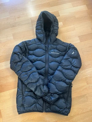 Svart dunjacka Peak Performance - Säljer en svart dunjacka från Peak Performance med huva och dragkedja framtill. Jackan har quiltad design och långärmade ärmar, perfekt för kalla dagar. Snygg och funktionell för dig som gillar stil och värme.