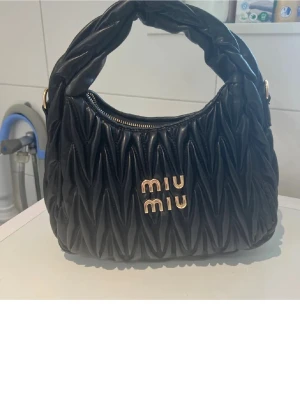 Svart quiltad handväska från Miu Miu - Säljer en svart quilt busig handväska från Miu Miu i skinn med guldiga detaljer och logga framtill. Väskan har ett vadderat handtag, dragkedja upptill och avtagbar axelrem. Perfekt för dig som vill ha en statementväska med lyxig känsla.