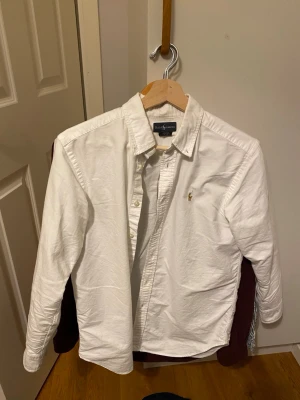 Vit skjorta från Ralph Lauren - Klassisk vit skjorta från Ralph Lauren med button-down krage och broderad logga på bröstet. Skjortan har långa ärmar och knäppning framtill. Perfekt till jeans eller chinos för en clean och stilren look. Kan gå ner i pris om det blir en snabb affär! Storlek 16 (xs/s)