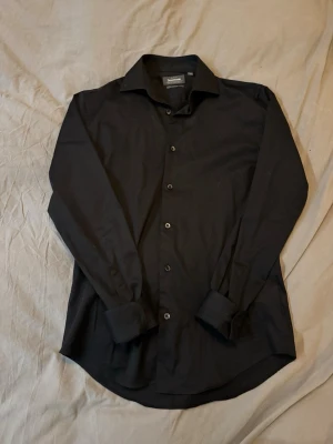 Svart skjorta  - Klassisk svart skjorta från Dressmann med lång ärm och knappar framtill. Slim fit passform. Skjortan har en stilren krage och breda manschetter, perfekt för dig som gillar en clean och enkel look. Tillverkad i mjuk bomull som känns skön mot huden.