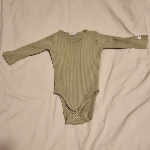 Ribbad grön body från Newbie - Säljer en ribbad grön body från Newbie med långa ärmar och rund hals. Bodyn har praktiska tryckknappar vid axeln och i grenen för enkel på- och avklädning.
