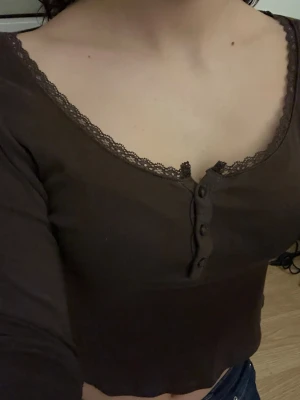 Brun långärmad topp med spets - Mysig brun långärmad topp från H&M med dekorativ spets runt halsringningen och knappar framtill. Modellen är v-ringad och har en slim passform som sitter snyggt på kroppen. Perfekt att styla med jeans eller kjol för en chill look.