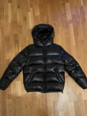 Svart pufferjacka från ZARA - Säljer en svart pufferjacka från ZARA i storlek M. Jackan har en stor huva, dragkedja framtill och två sidofickor. Materialet är glansigt och känns riktigt modernt, perfekt för kalla dagar. Passformen är normal och jackan är vadderad för extra värme.
