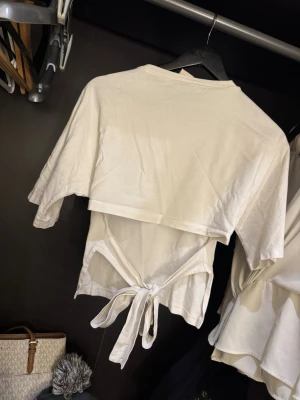 Vit topp med knytband - Snygg vit topp i bomull med korta ärmar och knytband i midjan. Toppen har en enkel och clean look med rund halsringning och öppen detalj baktill där banden knyts. Perfekt för en chill och trendig stil.