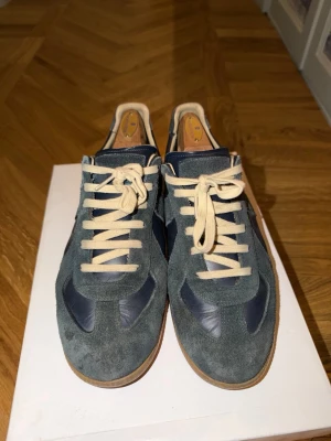 Maison Margiela sneakers i mocka och skinn - Säljer ett par Maison Margiela Gats i mörkblå mocka och skinn med beige snörning och gummisula. Klassisk retrostil med rund tå och snygga kontraster mellan material och färger. Perfekta för dig som gillar stilrena men unika sneakers. Använt flitigt så inte i nyskick. Har slitningar vid hälen och tecken på användning men fortfarande lika snygga som nyköpta.