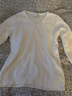 Vit stickad tröja med v-ringning från Vila - Snygg vit stickad tröja från Vila med v-ringad hals och långa ärmar. Tröjan har ribbad nederkant som ger en schysst detalj och är perfekt för lager-på-lager. Enkel och stilren design som funkar till många olika outfits.