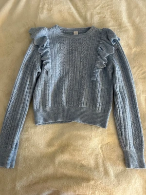 Ljusblå stickad tröja med volanger Lindex - Supersöt ljusblå stickad tröja från Lindex med snygga volangdetaljer på axlarna. Tröjan har ett diskret mönster, rund hals och långa ärmar. Perfekt för dig som gillar en trendig och mysig look. Passar dig som vill ha något extra till garderoben.