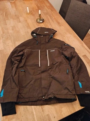 Brun skidjacka Peak Performance S - Brun skidjacka från Peak Performance med blå detaljer och huva. Jackan har flera dragkedjeförsedda fickor, justerbar nederkant och är tillverkad i vattenavvisande syntetmaterial. 