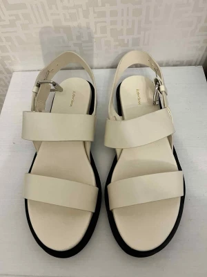 Beige sandaler med breda remmar - Snygga beige sandaler från A Better Story med breda remmar och silverfärgat spänne. Platt svart sula och öppen tå ger en clean och modern vibe. Perfekta för varma dagar och enkel att matcha med olika outfits.