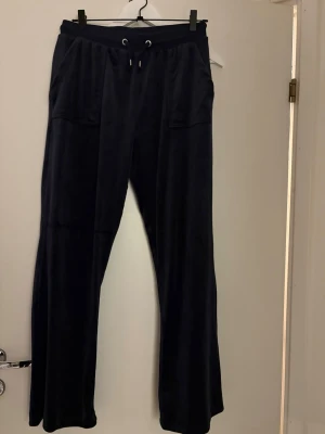 Marinblåa  mjukisbyxor från Juicy Couture - Marinblå mjukisbyxor från Juicy Couture med bred resår och snörning i midjan. Byxorna har stora fickor fram och bak med silverfärgade detaljer. Materialet är mjukt och sammetliknande, och modellen är loose med raka ben. Storlek 15/16 - kidsbrandstore