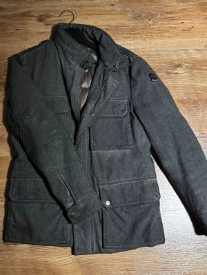 Mörkgrå parkajacka från Moncler - Parkajacka från Moncler i mörkgrått med quiltad insida och flera fickor framtill. Jackan har långärmade ärmar, hög krage och snygga knappdetaljer. Yttermaterialet är ull och insidan är fodrad med polyester för extra värme. Perfekt för kalla dagar. Sitter tajt mer S en M.