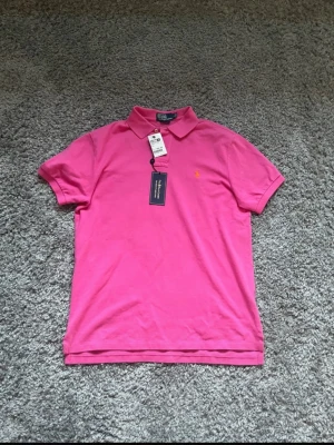 Rosa pikétröja från Polo Ralph Lauren - Säljer en snygg rosa pikétröja från Polo Ralph Lauren i storlek L. Tröjan har klassisk krage, korta ärmar och två knappar vid halsen. Materialet är bomull och den har en gul broderad logga på bröstet. Perfekt för dig som gillar stilrena och färgglada plagg.