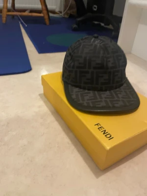 Svart Fendi keps med FF-mönster - Säljer en svart Fendi keps med ikoniskt FF-mönster över hela kepsen. Kepsen har böjd skärm med läderdetalj och justerbar rem baktill med Fendi-logga. Materialet är polyester och bomull, vilket gör den både snygg och bekväm.äkta kommer med allt 