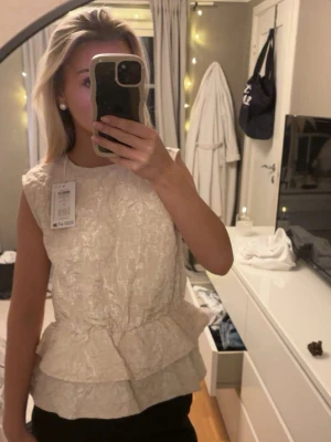 Beige blus med volang och struktur - Säljer en beige ärmlös blus med vacker struktur och dubbla volanglager nedtill. Blusen har rund halsringning och en lätt skimrande yta som ger en lyxig känsla. Perfekt för dig som gillar feminina detaljer och vill sticka ut lite extra.