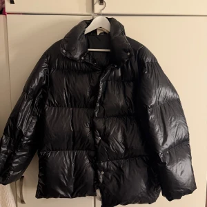 Svart pufferjacka med hög krage - Säljer en svart pufferjacka från Arket med glansig finish och hög krage. Jackan har breda, quiltade sektioner och stängs med knappar framtill. Perfekt för kalla dagar när du vill ha en cool och oversized look.