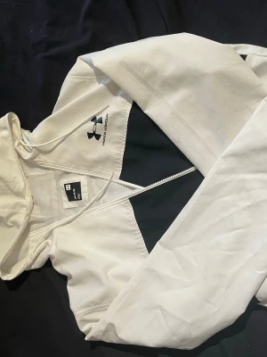 Vit och svart vindjacka Under Armour - Säljer en snygg vit och svart vindjacka från Under Armour i storlek M. Jackan har huva med dragsko, hel dragkedja framtill och meshfoder för extra komfort. Perfekt för dig som vill ha en sportig look och gillar funktionella detaljer.