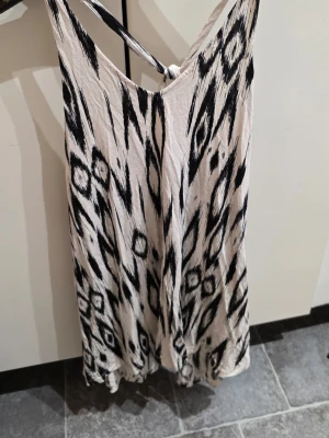 Vit och svart mönstrad klänning H&M XS - Säljer en snygg kortklänning från H&M i storlek XS. Klänningen är vit med svart grafiskt mönster, har smala axelband med knyt i ryggen och är tillverkad i mjuk bomull. Perfekt för dig som gillar unik stil och bekväma material.