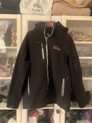 Svart vindjacka från Toyota - Säljer en svart vindjacka från Toyota Material Handling med huva och vita dragkedjor. Jackan har flera fickor med dragkedja, justerbara ärmslut och en sportig look. Perfekt för dig som vill ha en funktionell och stilren jacka till vardags.