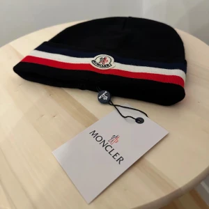 Moncler svart mössa med ränder - Svart mössa Moncler med klassiska ränder i marinblått, vitt och rött längs kanten. Märkets logga är broderad framtill. Mössan har en stilren och enkel form, perfekt för dig som gillar exklusiva accessoarer. Materialet är mjukt och känns lyxigt.