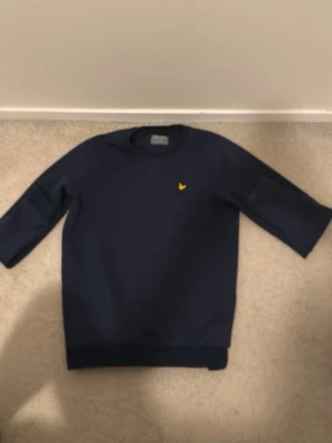 Marinblå sweatshirt från Lyle & Scott - Snygg marinblå sweatshirt från Lyle & Scott med klassisk rund hals och gul broderad logga på bröstet. Tröjan har långärmad design och ribbade muddar vid ärmslut och nederkant. Perfekt för en chill och stilren look.