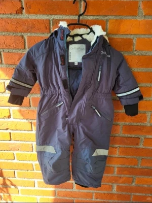 Grå snöoverall från On The Peak storlek 80 - Säljer ett par lila snökläderr med reflexdetaljer och dragkedjor från On The Peak. Byxorna har fuskpäls i luvan, mjukt vitt foder och flera praktiska fickor med dragkedja. Perfekt för kalla dagar med AquaTex air vent-material som andas.
