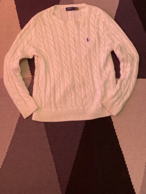 Polo Ralph Lauren - Säljer en vit kabelstickad tröja från Polo Ralph Lauren i storlek S. Tröjan är tillverkad i 100% bomull och har ett klassiskt kabelmönster samt rund hals. Liten blå logga broderad på bröstet och ribbade muddar vid ärmslut och nederkant.
