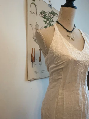 Vit broderad sommarklänning i linne - Säljer en vit klänning i linne med broderade blommor på bröstet. Klänningen har v-ringad hals, ärmlös design och figurnära passform. Perfekt för varma dagar och har en fräsch, enkel look med fina detaljer.