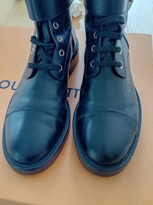 Svarta boots från Louis Vuitton - Säljer ett par svarta boots från Louis Vuitton med guldfärgade LV-detaljer vid ankeln. Skorna har snörning med svarta skosnören och guldfärgade öljetter. Tillverkade i slätt skinn med rund tå och platt sula. Perfekt för dig som gillar lyxiga och stilrena skor.Storlek 39