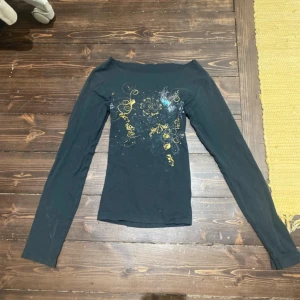 Svart långärmad topp med guldmönster - Säljer en svart långärmad topp med coolt guldigt och blått mönster framtill. Toppen har en smal passform och är gjord i mjukt material, perfekt att styla med jeans eller kjol. Unik design med konstnärliga detaljer som verkligen sticker ut.