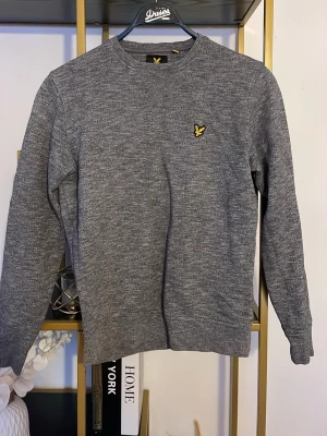 Grå tröja från Lyle & Scott - Säljer en stilren grå tröja från Lyle & Scott med klassisk rund hals och långärmad design. Tröjan har ett litet gult fågelbroderi på bröstet och är tillverkad i mjuk bomullsmix. Perfekt för dig som gillar enkel och snygg stil.