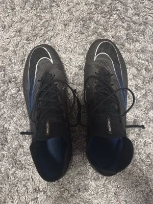 Nike Zoom Mercurial Superfly 9 Elite Ag pro Shadow Pack - Säljer ett par svarta Nike Mercurial Air Zoom fotbollsskor med blå och vita detaljer. Skorna har snörning, strumpliknande skaft och en lätt, syntetisk ovandel. Sulan har dobbar för bra grepp på planen och designen är riktigt sportig och modern.