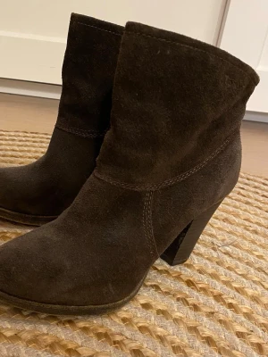 Bruna mockaboots med klack - Snygga bruna boots i mjuk mocka med stabil klack. Perfekta för dig som gillar en stilren look med lite höjd. Bootsens enkla design gör dem lätta att matcha med olika outfits och de har en klassisk form som aldrig går ur tiden.