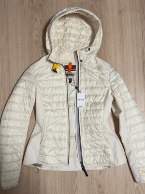 Vit pufferjacka från Parajumpers XS - Snygg vit pufferjacka från Parajumpers i storlek XS. Jackan har huva, dragkedja framtill och coola detaljer som gul och orange rem vid halsen. Materialet är polyester och jackan har quiltad design. Perfekt för dig som vill ha en lätt och stilren jacka.
