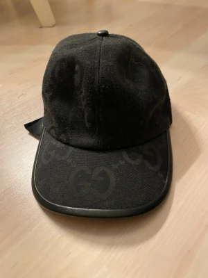 Svart  keps med GG-mönster - Säljer en svart keps från  med det klassiska GG-mönstret ton-i-ton över hela kepsen. Kepsen har böjd skärm med svart kant och är tillverkad i ett slitstarkt tygmaterial. Insidan har detaljerad märkning. Perfekt för dig som gillar exklusiva accessoarer.
