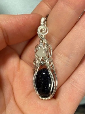 Handgjord wire wrap-hänge med blå glittrig sten - Unikt handgjort hänge i silverfärgad wire med en oval, mörkblå glittrande sten och en mindre rund, ljus sten upptill. Snygga snirkliga detaljer i metallen ger ett magiskt och elegant intryck. Perfekt för dig som gillar boho eller mystisk stil.