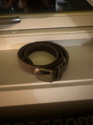 Brunt flätat bälte med metallspänne - Snyggt brunt bälte med flätad design och ett stilrent metallspänne i silverfärg. Bältet är gjort av textilmaterial och har läderdetaljer vid spännet. Perfekt för att ge outfiten en extra touch.